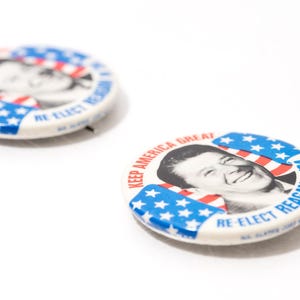 May include: Two vintage campaign badges on a white surface. The badges feature a portrait of a smiling man, with red, white, and blue stripes and stars. Text reads "KEEP AMERICA GREAT" and "RE-ELECT REAGAN IN '84."
