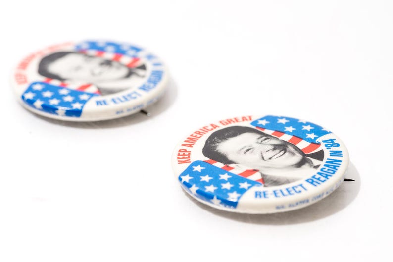 May include: Two vintage campaign badges on a white surface. The badges feature a portrait of a smiling man, with red, white, and blue stripes and stars. Text reads "KEEP AMERICA GREAT" and "RE-ELECT REAGAN IN '84."