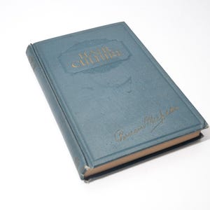 May include: A vintage book titled "Hair Culture" in a teal cover with gold lettering. The book has a worn appearance, suggesting age. The author's signature is also visible in gold on the cover.