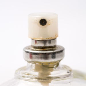 May include: Close-up of a perfume bottle's sprayer. The sprayer has a cream-colored cap with a small black nozzle. The metal collar shows signs of wear. The clear glass bottle is partially visible.