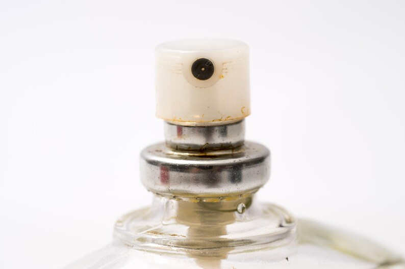 May include: Close-up of a perfume bottle's sprayer. The sprayer has a cream-colored cap with a small black nozzle. The metal collar shows signs of wear. The clear glass bottle is partially visible.