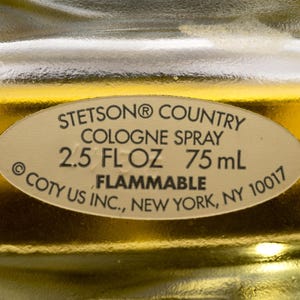 May include: Close-up of a cologne bottle label. The label reads "STETSON&reg; COUNTRY COLOGNE SPRAY 2.5 FL OZ 75 mL FLAMMABLE &copy; COTY US INC., NEW YORK, NY 10017". The background is a golden color.