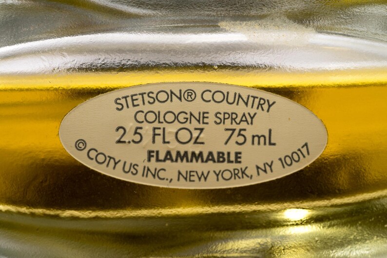 May include: Close-up of a cologne bottle label. The label reads "STETSON&reg; COUNTRY COLOGNE SPRAY 2.5 FL OZ 75 mL FLAMMABLE &copy; COTY US INC., NEW YORK, NY 10017". The background is a golden color.