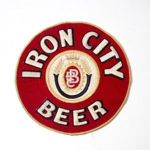 May include: A circular embroidered patch featuring a red background with the text IRON CITY BEER in white letters with black outlines. The center displays a white circle with a gold wheat design, a crown, and a red oval containing the letters BC.