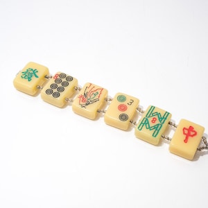 May include: A vintage bracelet made of six rectangular mahjong tiles. The tiles are cream-colored with various symbols and numbers in green, red, and black. The tiles are connected by small silver-colored links.