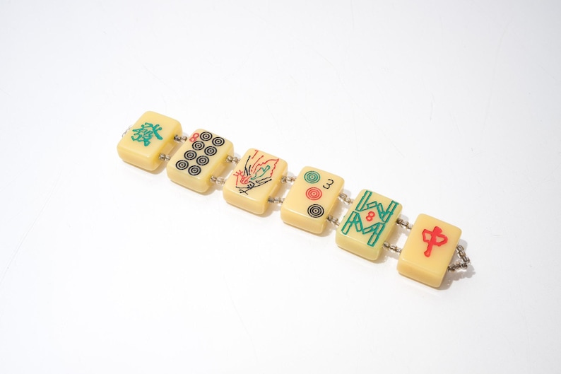 May include: A vintage bracelet made of six rectangular mahjong tiles. The tiles are cream-colored with various symbols and numbers in green, red, and black. The tiles are connected by small silver-colored links.
