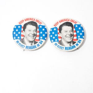 May include: Two circular campaign badges. Each badge displays a portrait of Ronald Reagan, with the text "KEEP AMERICA GREAT" and "RE-ELECT REAGAN IN '84" in red and blue, with an American flag design.