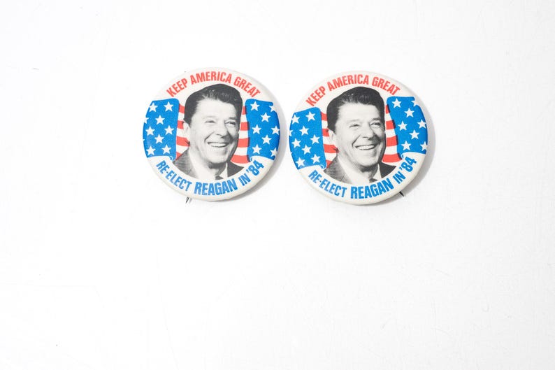 May include: Two circular campaign badges. Each badge displays a portrait of Ronald Reagan, with the text "KEEP AMERICA GREAT" and "RE-ELECT REAGAN IN '84" in red and blue, with an American flag design.