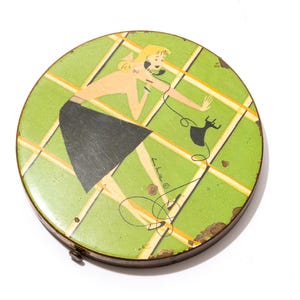 Op de afbeelding: Vintage ronde metalen compactpoederdoos met een groen en geel rasterpatroon. Het deksel toont een cartoonillustratie van een vrouw aan de telefoon. De compactpoederdoos heeft een bruine metalen rand en vertoont tekenen van ouderdom.