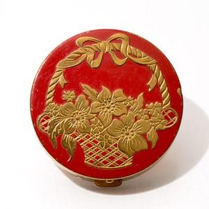 Op de afbeelding: Een ronde, rode compactpoederdoos met een goudkleurig bloemenmandontwerp op het deksel. De mand is gevuld met bloemen en heeft een strik aan de bovenkant. De compactpoederdoos heeft een goudkleurige metalen rand en sluiting.