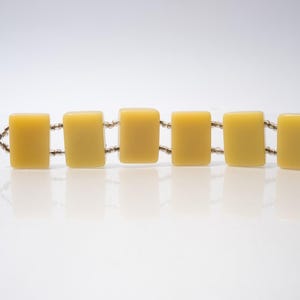 May include: A vintage bracelet featuring six rectangular, yellow-toned beads connected by small, silver-colored links. The bracelet is displayed on a white surface, showcasing its simple yet elegant design. The beads have a smooth, polished appearance.