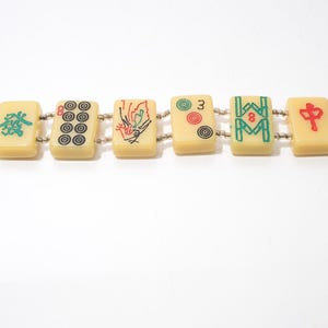 May include: A bracelet featuring six rectangular tiles, each displaying unique symbols and characters in red, green, and black. The tiles are connected by small, silver-colored beads. The bracelet is set against a white background.