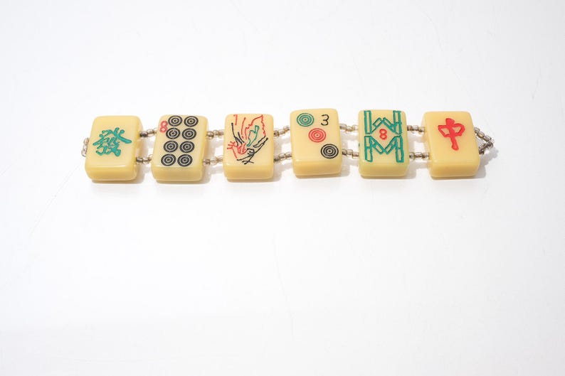 May include: A bracelet featuring six rectangular tiles, each displaying unique symbols and characters in red, green, and black. The tiles are connected by small, silver-colored beads. The bracelet is set against a white background.