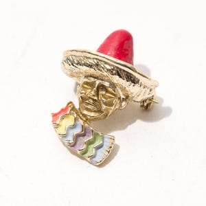May include: Gold-toned brooch depicting a man wearing a red sombrero and holding a colourful fan. The man's face is detailed and the fan has a zig-zag pattern.