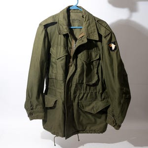 May include: An olive green field jacket featuring a collared neckline, a front zipper closure, and four large button-flap pockets. The left sleeve displays a patch with an eagle emblem. The interior collar area contains printed text labels.