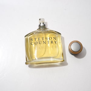 May include: A clear glass bottle of Stetson Country cologne with a silver and brown cap. The bottle is filled with a light yellow liquid and the words "STETSON COUNTRY" are printed on the front in black.