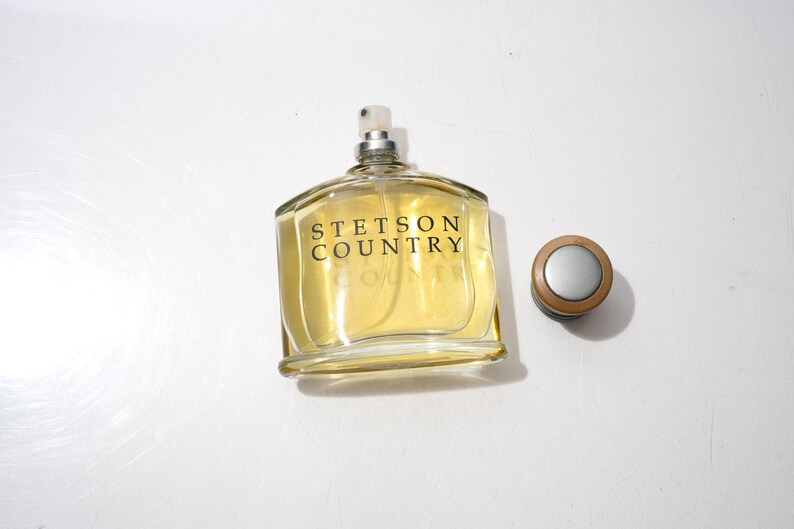 May include: A clear glass bottle of Stetson Country cologne with a silver and brown cap. The bottle is filled with a light yellow liquid and the words "STETSON COUNTRY" are printed on the front in black.