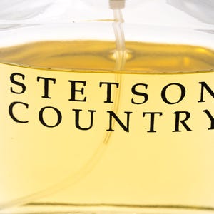 May include: Close-up of a clear glass bottle filled with a light amber liquid. The words "STETSON COUNTRY" are printed in black, bold letters on the front of the bottle. A thin, clear spray nozzle extends from the top.