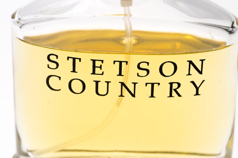 May include: Close-up of a clear glass bottle filled with a light amber liquid. The words "STETSON COUNTRY" are printed in black, bold letters on the front of the bottle. A thin, clear spray nozzle extends from the top.