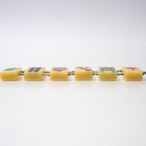 May include: A bracelet featuring six yellow, square tiles, each adorned with colorful symbols. The tiles are connected by small metal links. The bracelet is set against a white background.