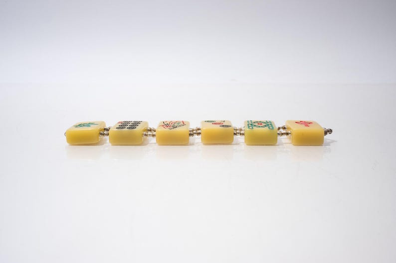 May include: A bracelet featuring six yellow, square tiles, each adorned with colorful symbols. The tiles are connected by small metal links. The bracelet is set against a white background.