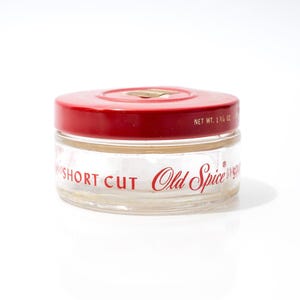 Puede incluir: Un tarro de cristal transparente con tapa roja. El tarro tiene las palabras "SHORT CUT" y "Old Spice" impresas en rojo. La tapa es plana y redonda. Peso neto: 49,6 gramos. Producto de cuidado personal.