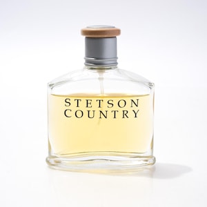 May include: Clear glass perfume bottle with a silver neck and wooden cap. The bottle contains a light yellow liquid and is labeled "STETSON COUNTRY" in black text. The bottle is on a white background.