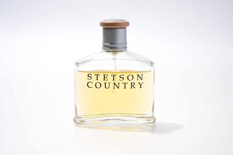 May include: Clear glass perfume bottle with a silver neck and wooden cap. The bottle contains a light yellow liquid and is labeled "STETSON COUNTRY" in black text. The bottle is on a white background.
