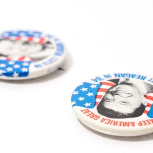 May include: Two vintage campaign badges. One badge features a black and white portrait of a man with the text "RE-ELECT REAGAN IN '84" and "KEEP AMERICA GREAT" in red and blue. The other badge is slightly out of focus.