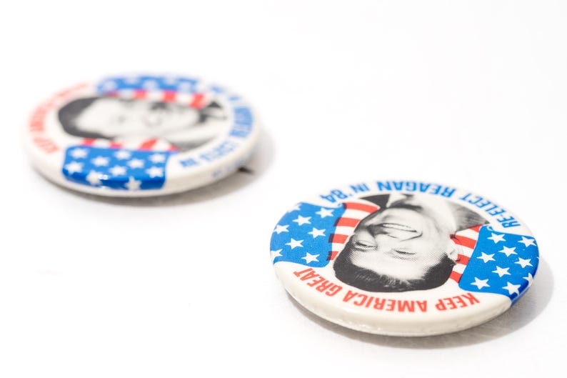 May include: Two vintage campaign badges. One badge features a black and white portrait of a man with the text "RE-ELECT REAGAN IN '84" and "KEEP AMERICA GREAT" in red and blue. The other badge is slightly out of focus.