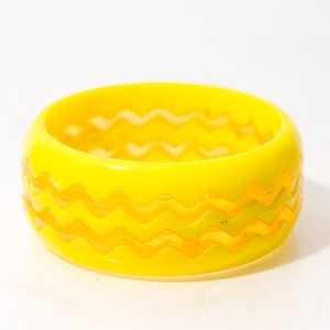 May include: A bright yellow bangle bracelet featuring a repeating horizontal zigzag pattern carved into the exterior surface. The circular accessory has a smooth, glossy finish and a consistent width throughout its entire rounded structure.
