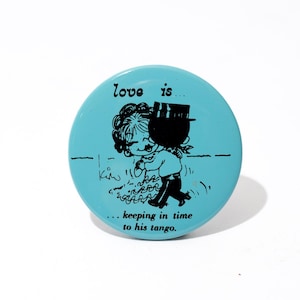 May include: A round, light blue button with a black and white illustration of a couple dancing. The text "love is... keeping in time to his tango" is printed on the button. The button is a vintage style.