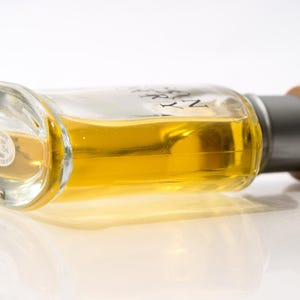 May include: A clear glass perfume bottle with a silver cap and a wooden top, filled with a golden-yellow liquid. The bottle is lying on its side, reflecting on a white surface.