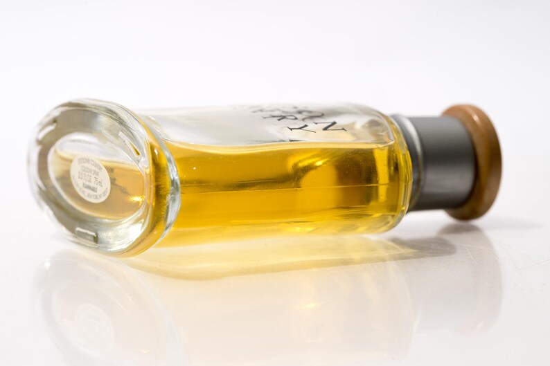 May include: A clear glass perfume bottle with a silver cap and a wooden top, filled with a golden-yellow liquid. The bottle is lying on its side, reflecting on a white surface.