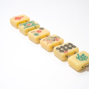 May include: A row of six ivory-colored Mahjong tiles with various symbols in red, green, and black. The tiles are connected with small metal loops. The background is plain white.