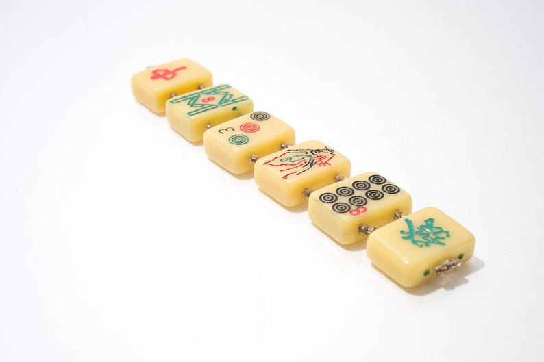 May include: A row of six ivory-colored Mahjong tiles with various symbols in red, green, and black. The tiles are connected with small metal loops. The background is plain white.