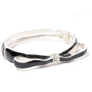 Puede incluir: Un brazalete de plata con un diseño de lazo de esmalte negro y un acento de cristal transparente.
