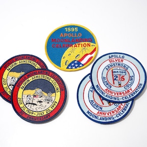 May include: A collection of embroidered patches commemorating the Apollo moon landing missions. The patches feature text, dates, and illustrations of the moon, astronauts, and the American flag. Colors include red, blue, yellow, and white.