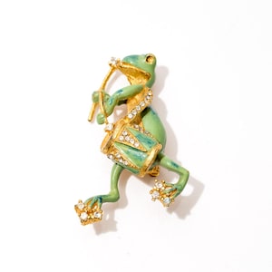 Puede incluir: Broche de rana caprichosa en metal dorado y esmalte. La rana verde toca un tambor adornado con pedrería. El broche tiene detalles de pedrería en los pies y la cabeza.