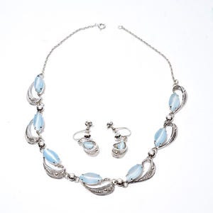 May include: A vintage jewelry set featuring a necklace and matching earrings. The necklace and earrings have a silver-tone metal setting with light blue, oval-shaped stones. The necklace has a delicate chain.