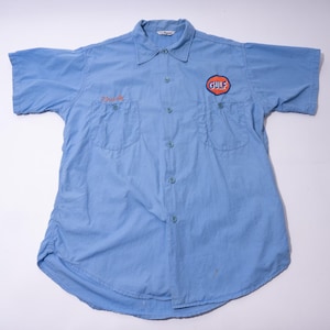 Puede incluir: Camisa de trabajo azul claro de manga corta con dos bolsillos en el pecho y cierre de botones. La camisa presenta el logotipo de Gulf en el pecho derecho y un nombre bordado sobre el bolsillo izquierdo. Tejido ligero.