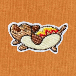 May include: Embroidered patch of a brown dog wearing a yellow hot dog costume with red and yellow mustard. The dog is lying on its back with its paws in the air.