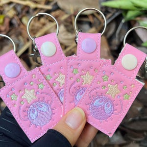 Puede incluir: Cuatro llaveros de cuero rosa con estrellas bordadas en morado y blanco y una criatura morada con ojos grandes. Cada llavero tiene un anillo de llave plateado y un botón rosa.