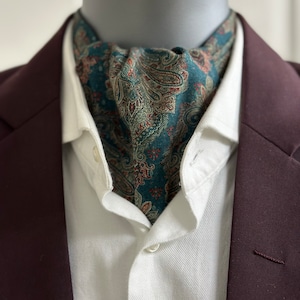 Può includere: Una camicia bianca con una cravatta a paisley verde acqua. La cravatta è annodata intorno al collo e la camicia è abbottonata al colletto. La camicia è indossata sotto un blazer bordeaux.