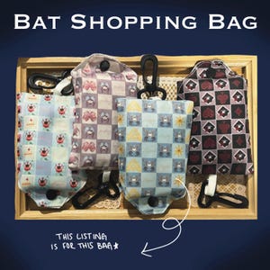 Puede incluir: Cuatro bolsas de compras con estampado de murci&eacute;lagos en una bandeja de madera. Las bolsas presentan varios dise&ntilde;os, incluyendo palos de cartas, lazos e ilustraciones de murci&eacute;lagos. Cada bolsa tiene un asa negra y un cierre a presi&oacute;n. El texto "BAT SHOPPING BAG" est&aacute; en la parte superior.