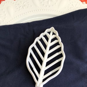 May include: White plastic leaf-shaped cookie cutter. The cutter has a detailed design with multiple lines and a rounded edge.