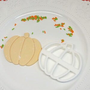 May include: A white ceramic pumpkin cookie cutter and a beige pumpkin cookie on a white plate with green and orange sprinkles.