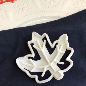 May include: White plastic leaf-shaped cookie cutter on a dark blue fabric background.