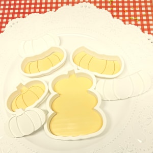 May include: A collection of white and yellow pumpkin-shaped cookie cutters on a white plate. The cutters are in various shapes and sizes, including a classic pumpkin, a long gourd, and a rounded pumpkin. The background features a red and white checkered pattern.
