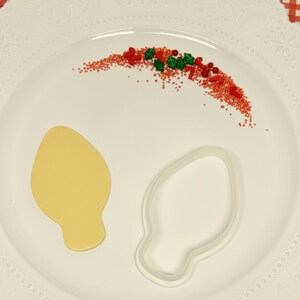 May include: A white plate with a yellow light bulb cookie cutter and a light bulb cookie. Red and green sprinkles are scattered on the plate.